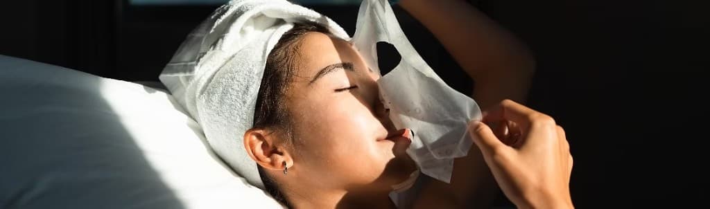 Woman removing a white sheet mask with a towel wrap, bright natural light